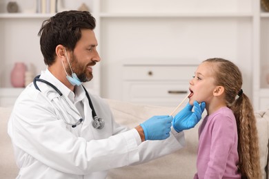 Photo of Smiling doctor examining girl`s oral cavity with tongue depressor indoors