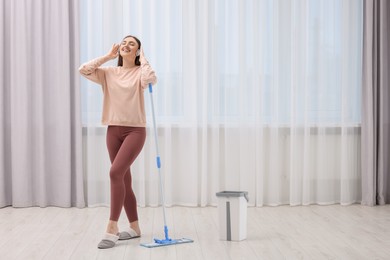 Photo of Enjoying cleaning. Happy woman in headphones listening music with mop indoors. Space for text