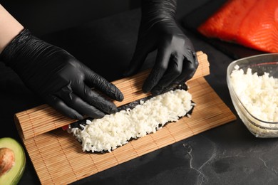 Photo of Chef in gloves wrapping sushi roll at dark textured table, closeup