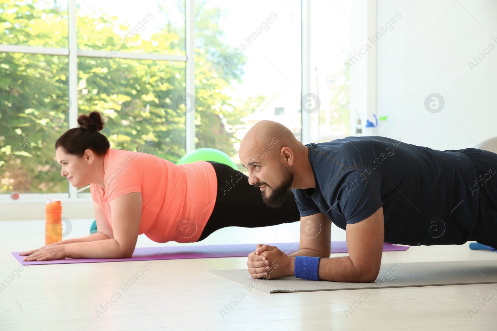 Photo of Overweight man and woman doing plank exercise on mats in gym