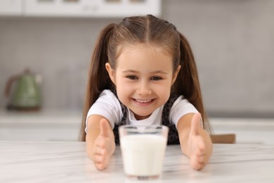 Photo of Cute girl reaching out for glass of milk at white table in kitchen