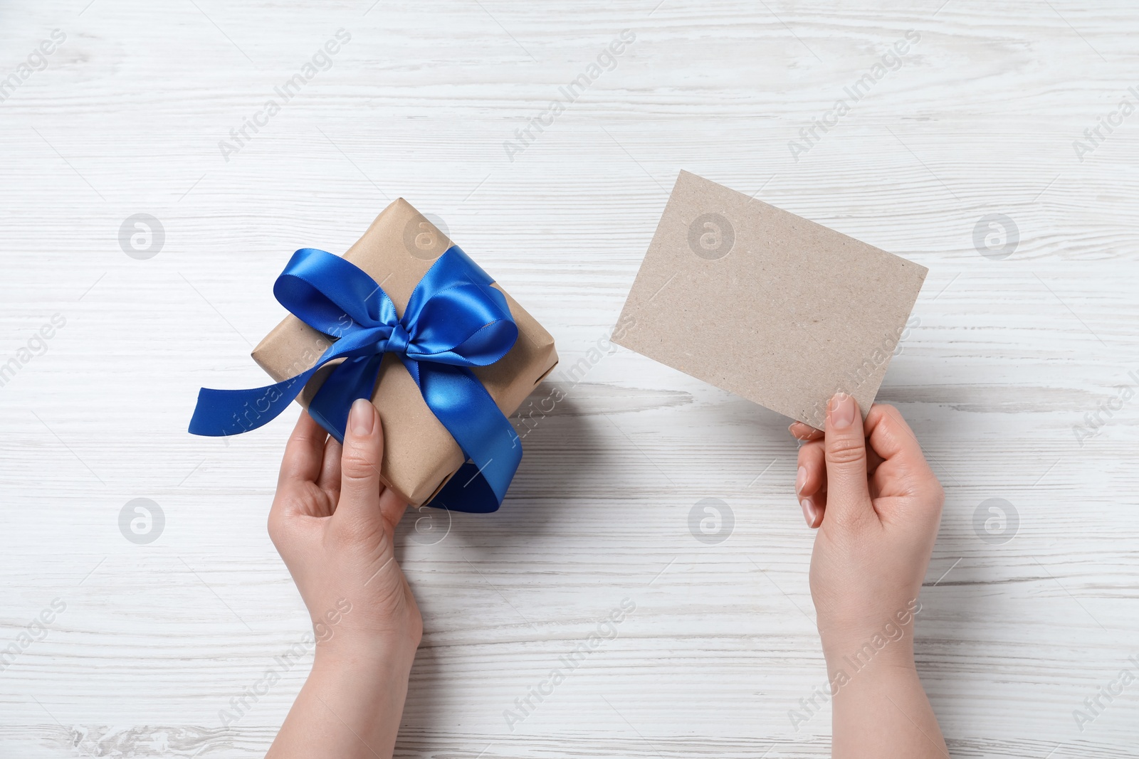 Photo of Woman holding gift box and blank card at white wooden table, top view. Space for text