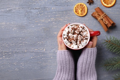 Woman holding cup of tasty cocoa with marshmallows on grey wooden table, top view. Space for text