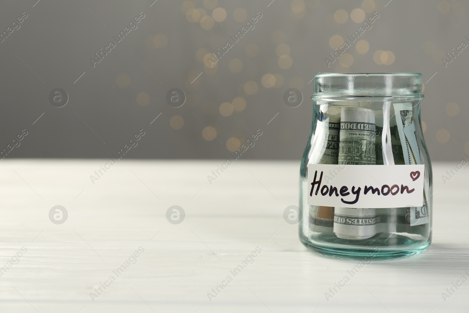 Photo of Glass jar with word Honeymoon and dollar banknotes on white wooden table, closeup. Space for text