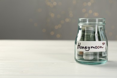 Glass jar with word Honeymoon and dollar banknotes on white wooden table, closeup. Space for text