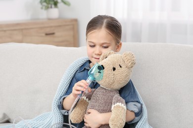Little girl with toy bear and nebulizer for inhalation on sofa at home
