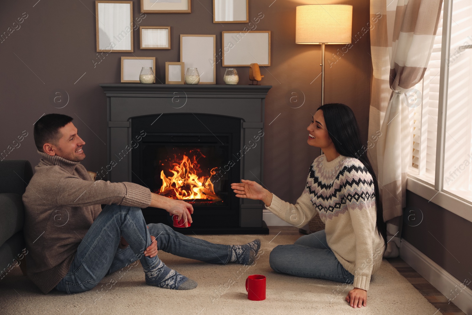 Photo of Lovely couple with cups of hot drink spending time together near fireplace at home