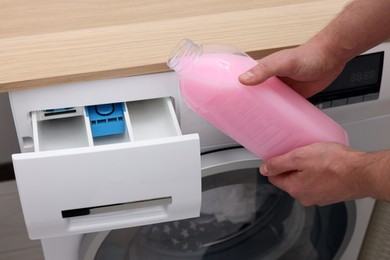 Photo of Man pouring fabric softener from bottle into washing machine indoors, closeup