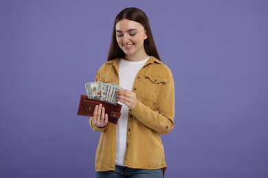 Happy woman holding wallet with dollar banknotes on purple background