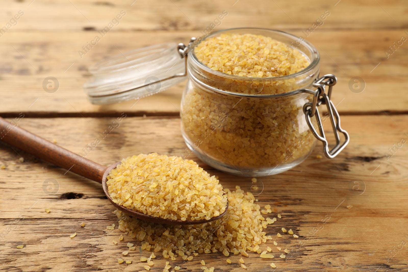 Photo of Glass jar and spoon with raw bulgur on wooden table, closeup