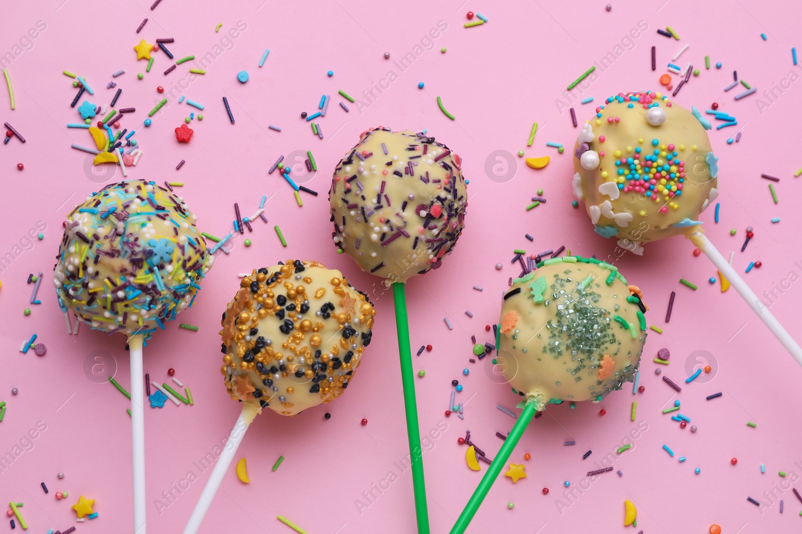 Photo of Delicious confectionery. Sweet cake pops and sprinkles on pale pink background, flat lay