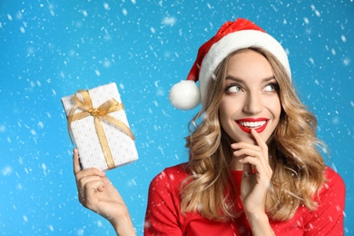 Happy young woman wearing Santa hat with Christmas gift on light blue background