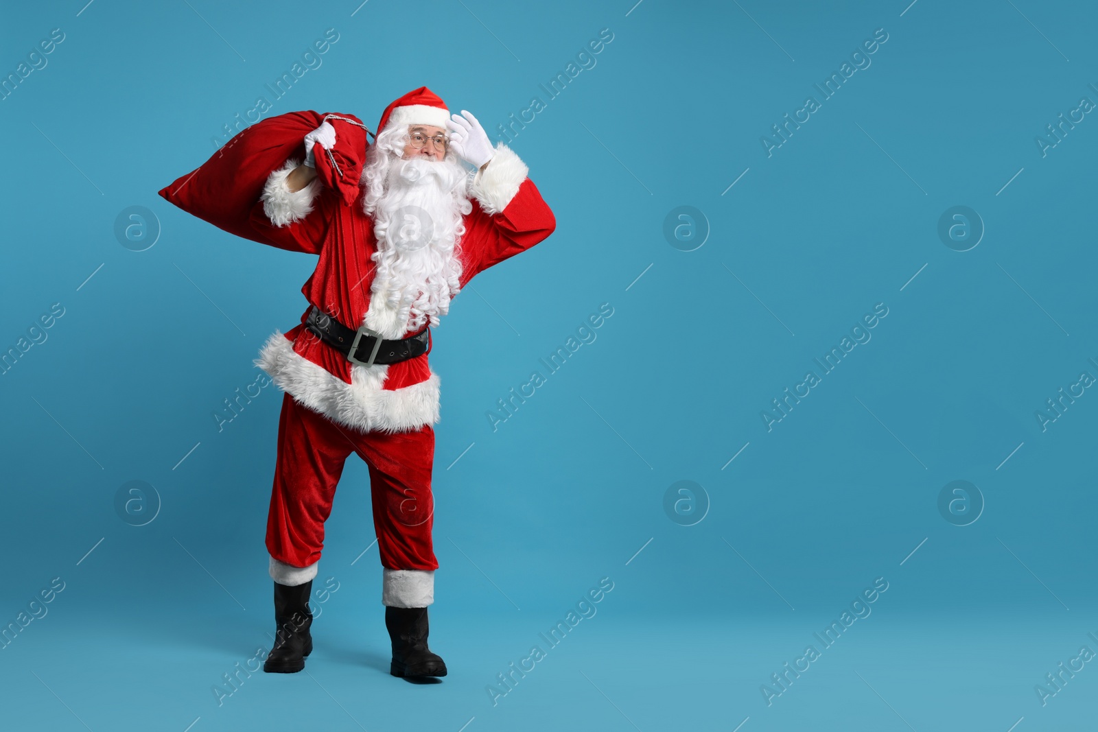 Photo of Santa Claus with bag of Christmas gifts posing on light blue background, space for text