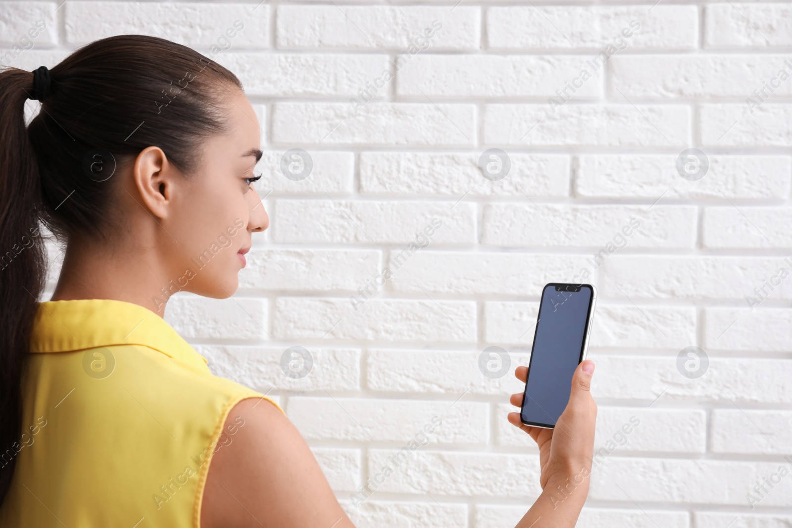 Photo of Young woman unlocking smartphone with facial scanner near white brick wall. Biometric verification