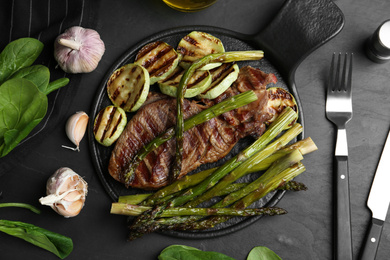 Tasty grilled meat served with asparagus and zucchini on black table, flat lay