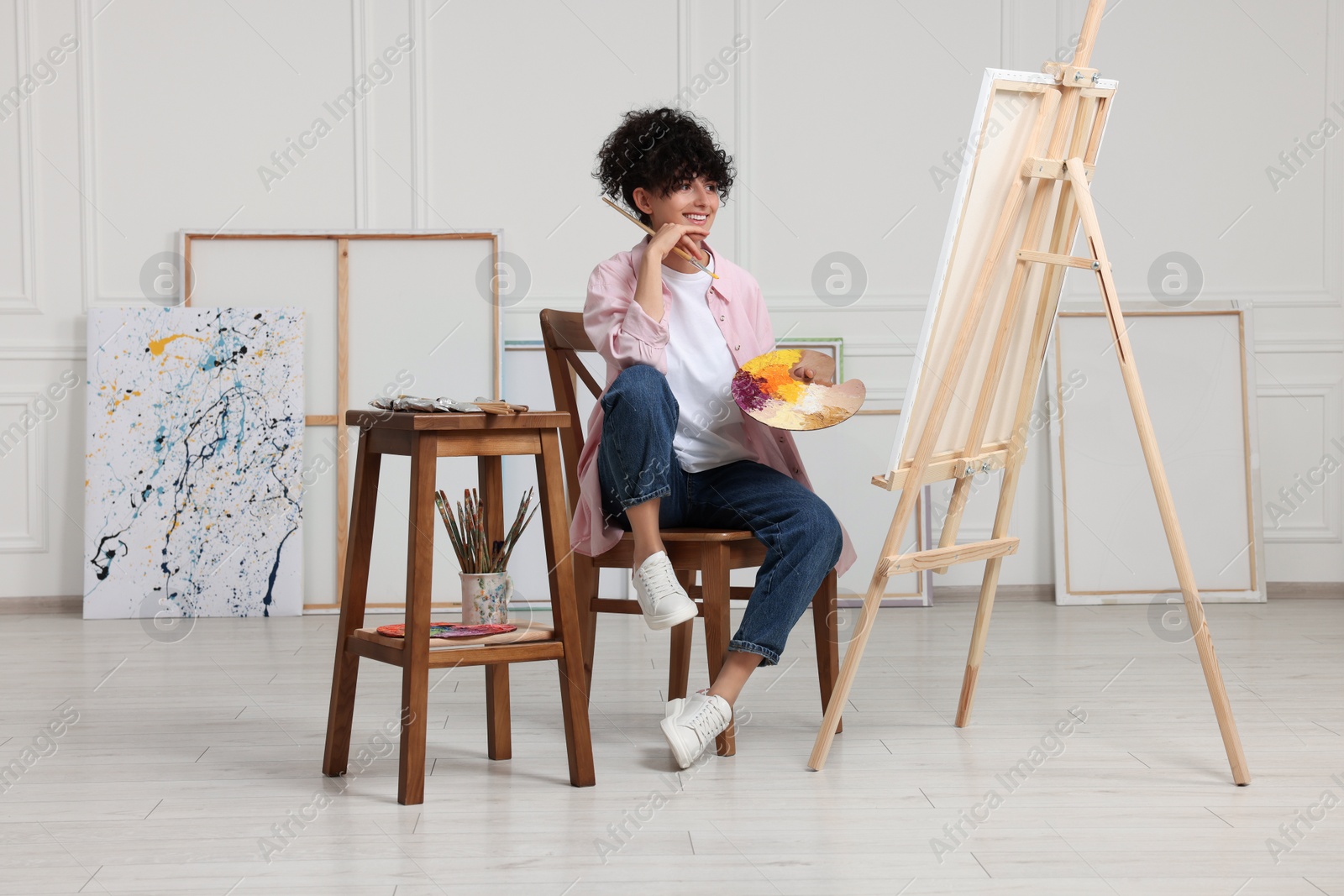 Photo of Young woman holding brush and artist`s palette near easel with canvas