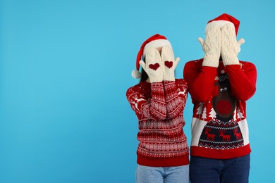 Photo of Young couple in Christmas sweaters and Santa hats covering faces with hands in knitted mittens on light blue background. Space for text