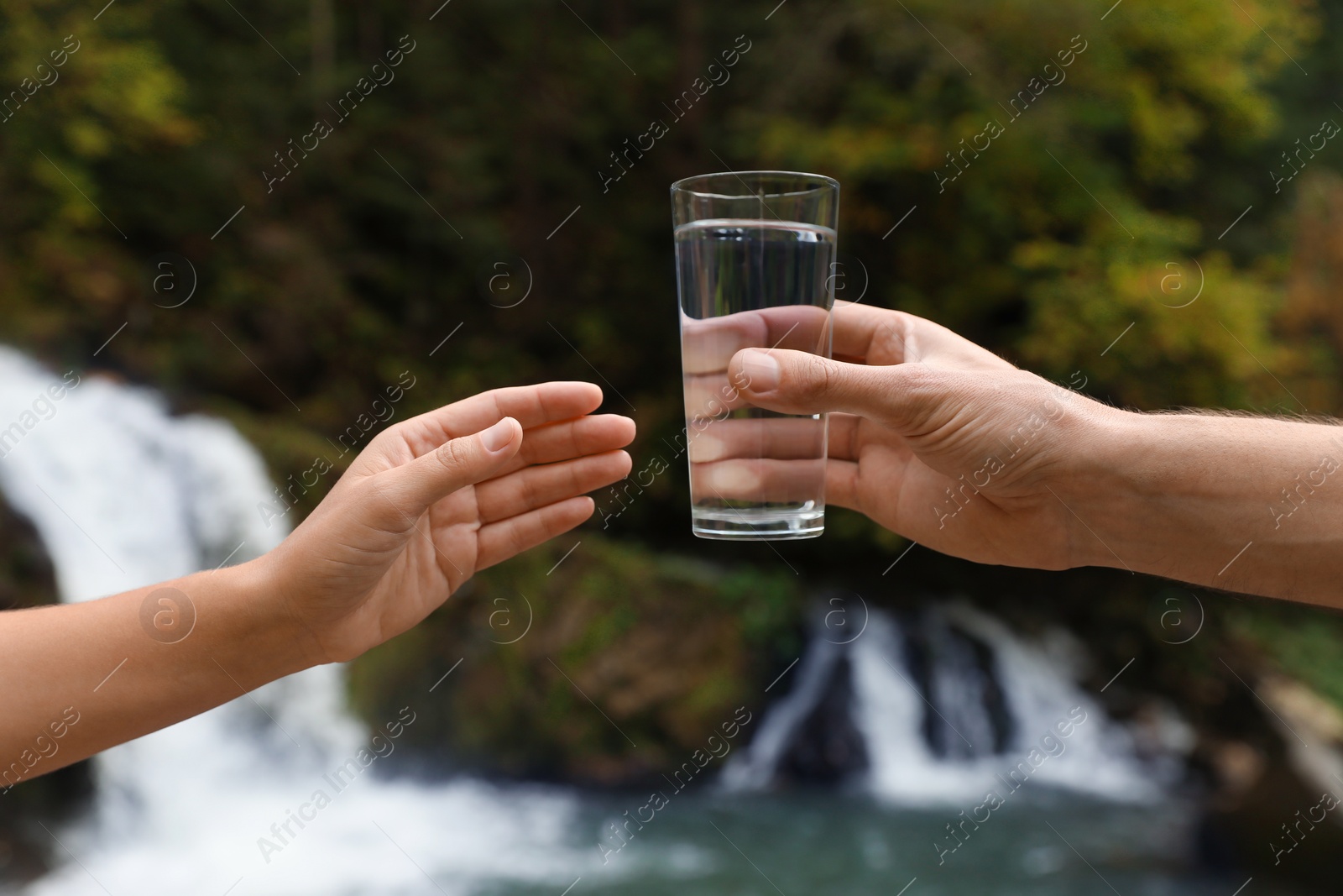 Photo of Man giving woman glass of fresh water near waterfall, closeup