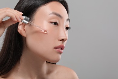 Photo of Beautiful young woman applying cosmetic serum onto her face on grey background, closeup. Space for text