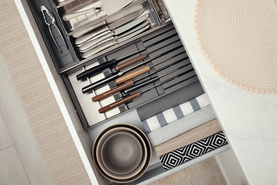 Open drawer of kitchen cabinet with different utensils, dishware and towels, top view