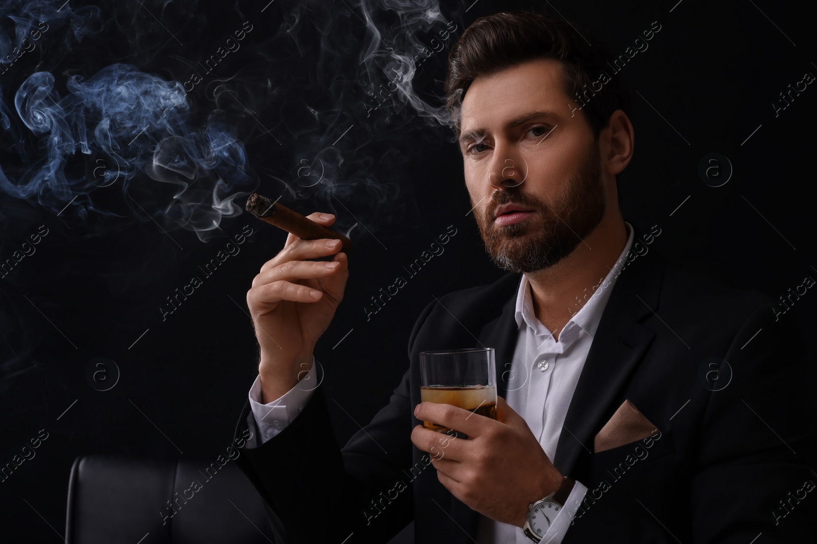 Photo of Handsome man in elegant suit with glass of whiskey smoking cigar on black background