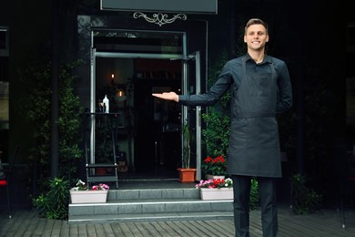 Happy young waiter in uniform near cafe, space for text