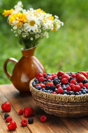 Photo of Wicker bowl with different fresh ripe berries and beautiful flowers on wooden table outdoors