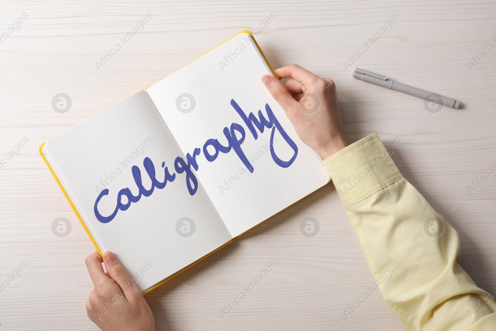 Image of Woman holding open notebook with written word Calligraphy at white wooden table, top view