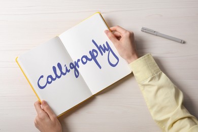 Image of Woman holding open notebook with written word Calligraphy at white wooden table, top view