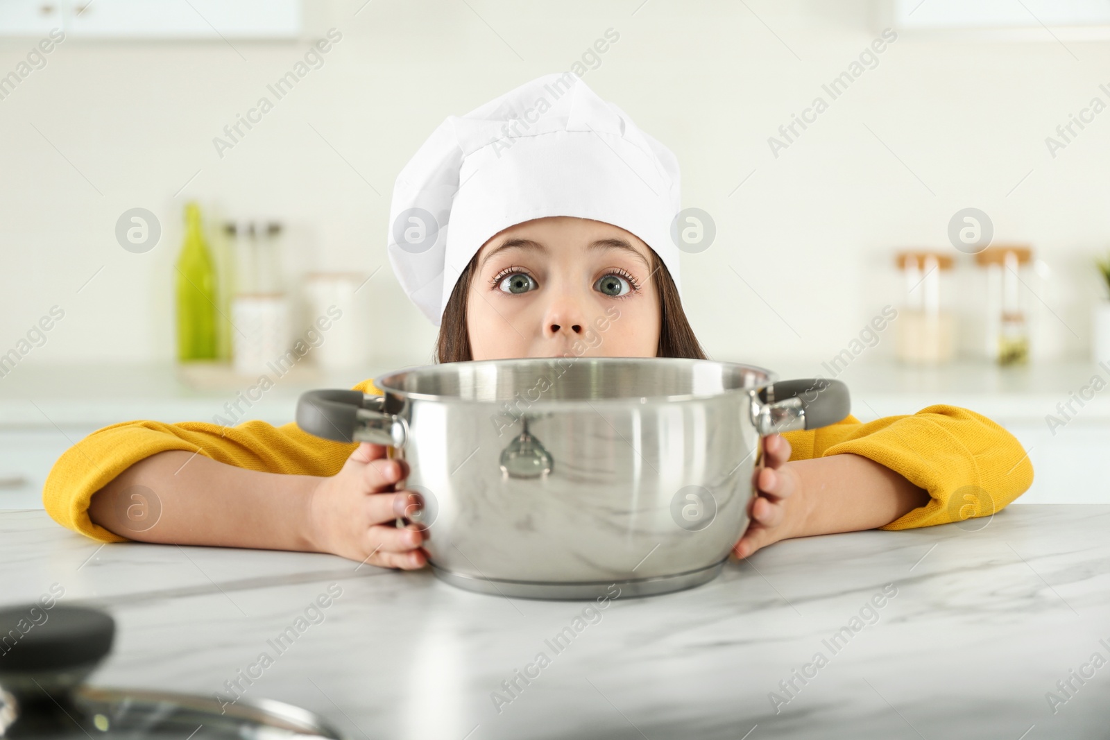 Photo of Surprised little girl wearing chef hat with pot in kitchen