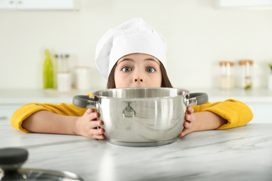 Surprised little girl wearing chef hat with pot in kitchen