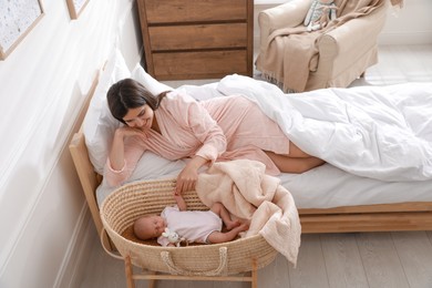Happy young mother resting near cradle with sleeping newborn baby at home