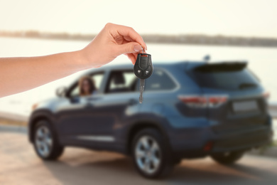 Car buying. Woman holding key against blurred automobile, closeup