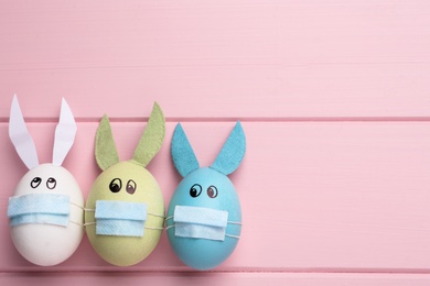 Photo of Dyed eggs with bunny ears in protective masks on pink wooden background, flat lay and space for text. Easter holiday during COVID-19 quarantine