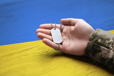 Image of Closeup of defender in camouflage uniform holding military ID tag over table. Wooden surface toned in colors of Ukrainian flag