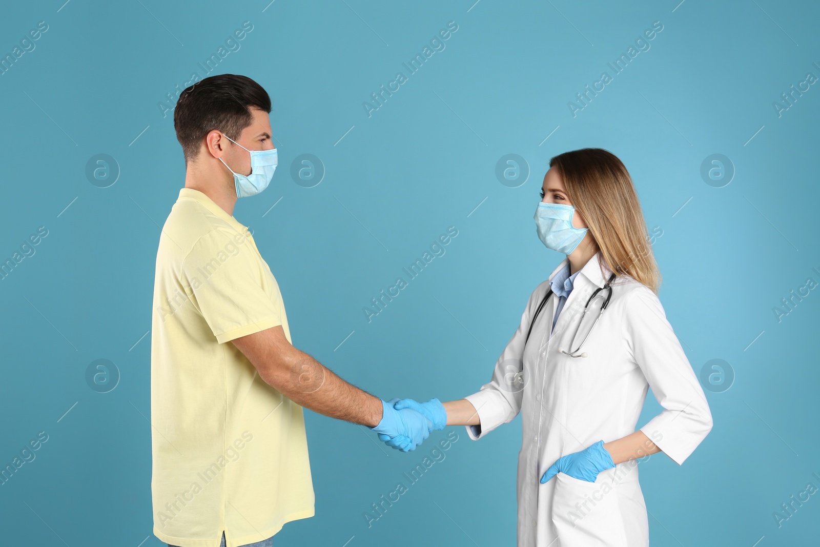 Photo of Doctor and patient in protective masks shaking hands on light blue background