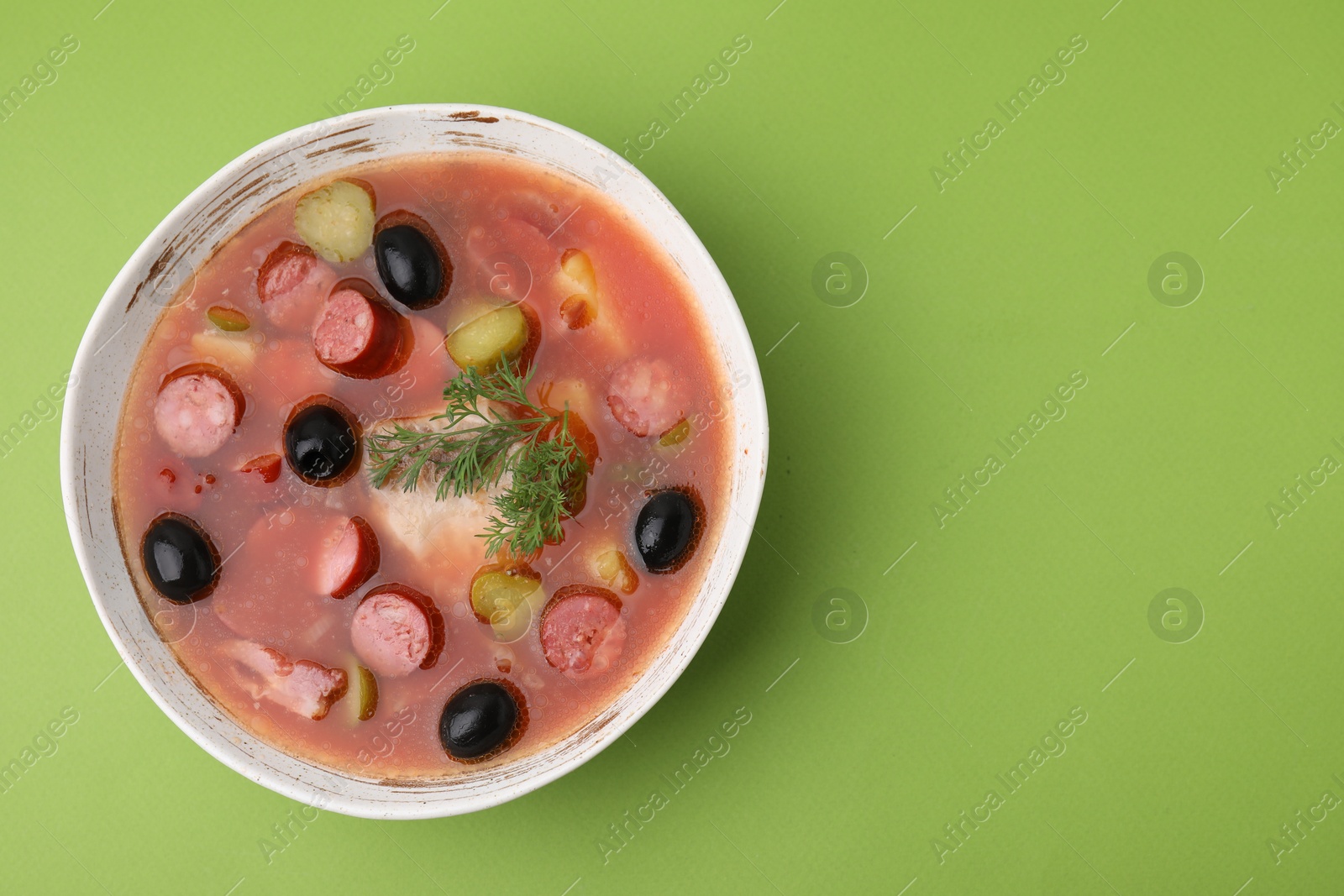 Photo of Meat solyanka soup with thin dry smoked sausages in bowl on light green background, top view. Space for text