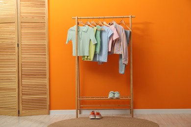 Photo of Rack with different stylish women's clothes, shoes and wooden folding screen near orange wall indoors