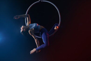 Young woman performing acrobatic element on aerial ring against dark background