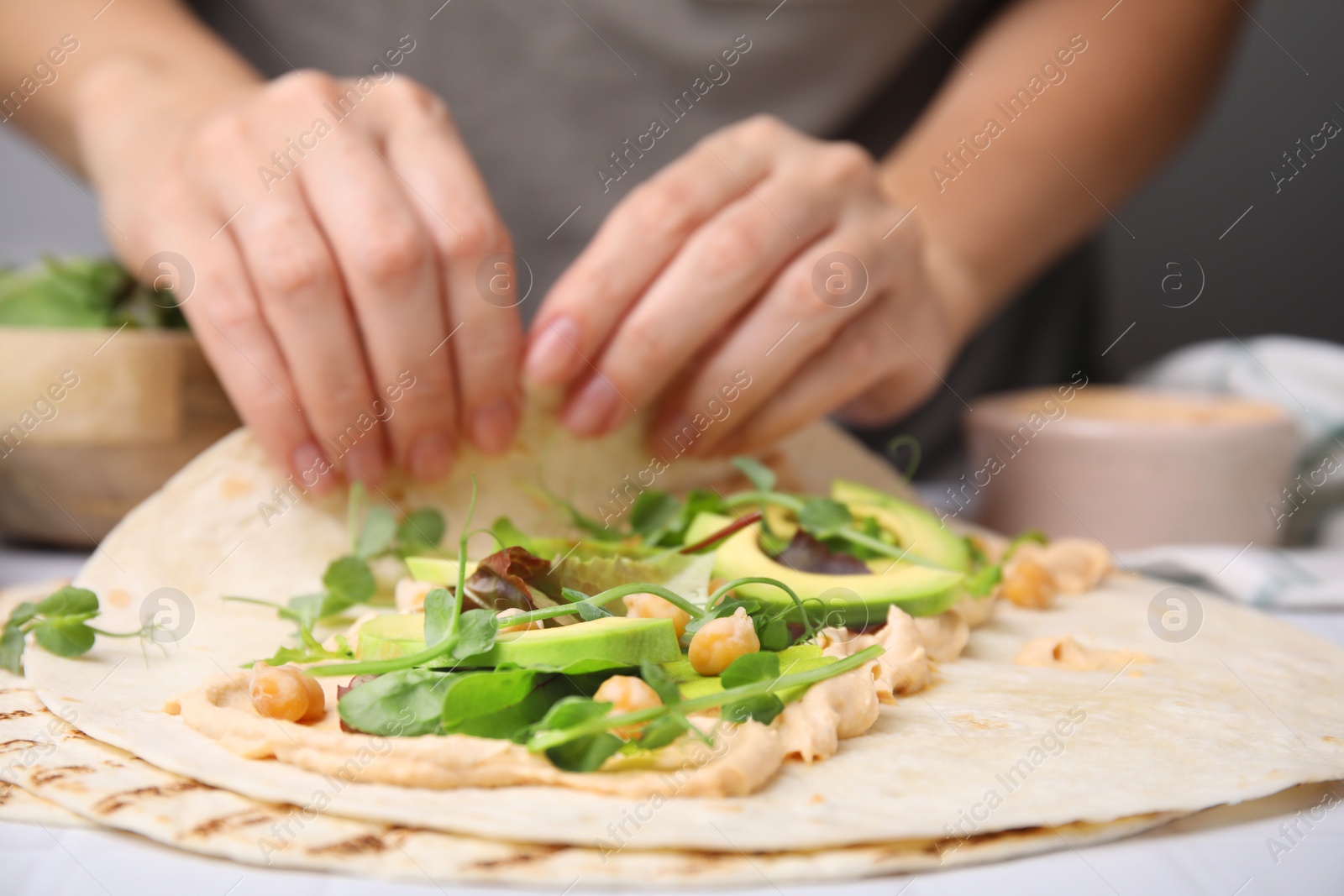 Photo of Woman making delicious hummus wrap with vegetables at table, closeup