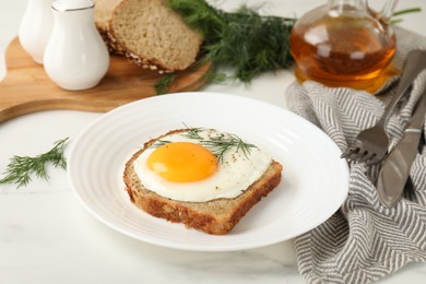 Plate with tasty fried egg, slice of bread and dill on white marble table