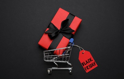 Image of Shopping cart, tag with phrase BLACK FRIDAY and gift box on dark background, flat lay