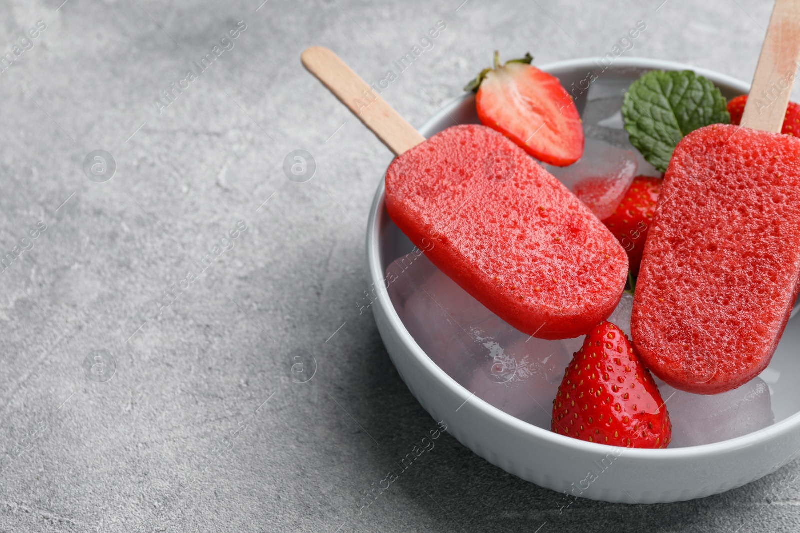 Photo of Bowl of tasty strawberry ice pops on grey table, space for text. Fruit popsicle