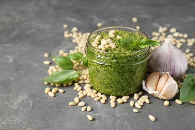 Photo of Jar of delicious pesto sauce and ingredients on grey table, closeup