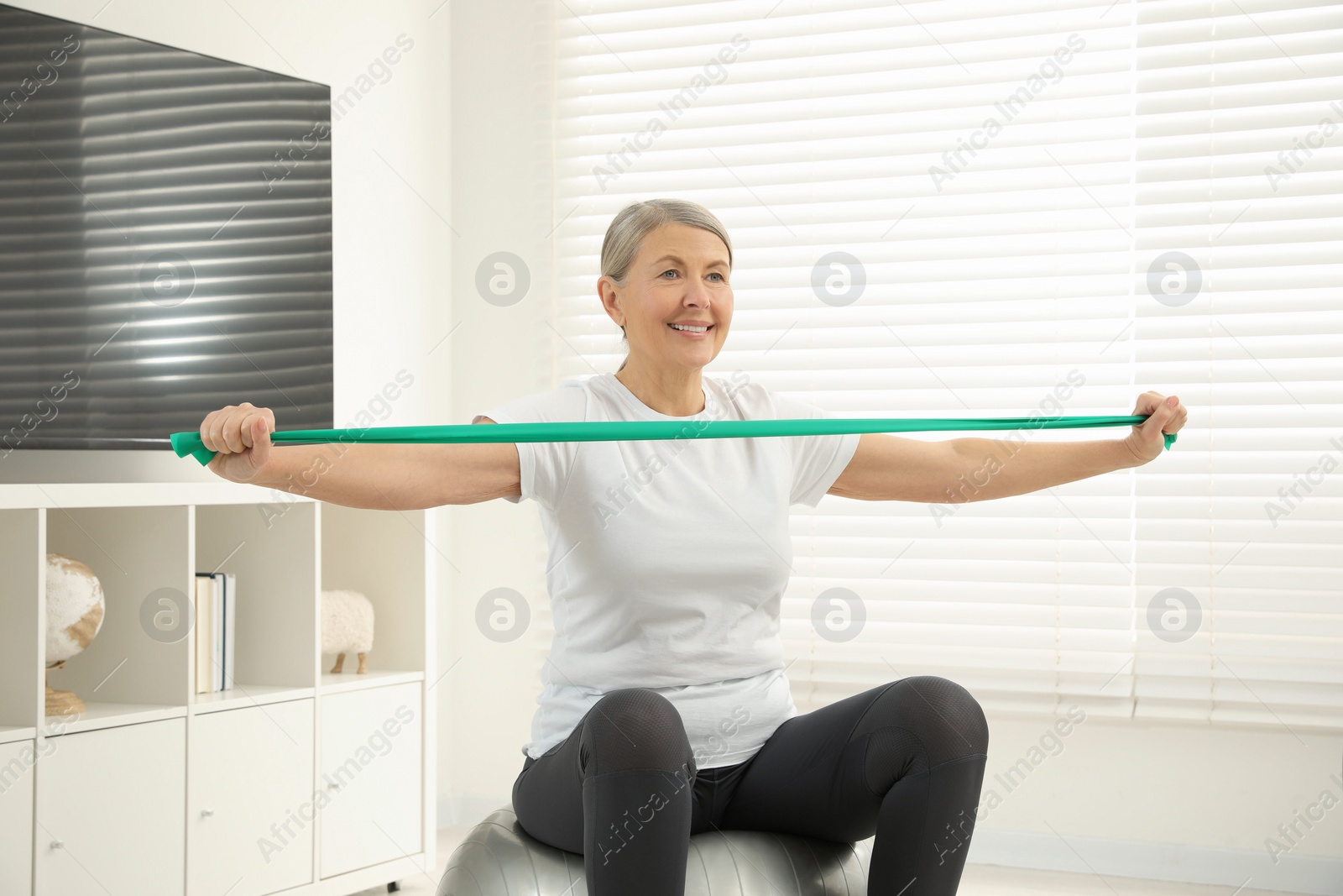 Photo of Senior woman doing exercise with elastic resistance band on fitness ball at home