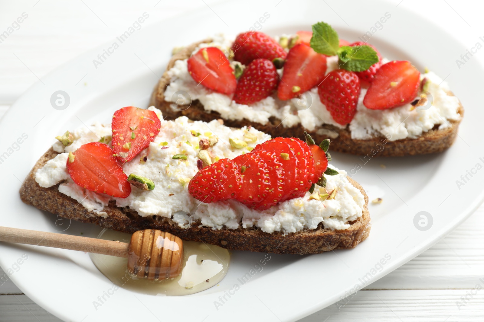 Photo of Delicious ricotta bruschettas with strawberry and pistachios served with honey on white table, closeup