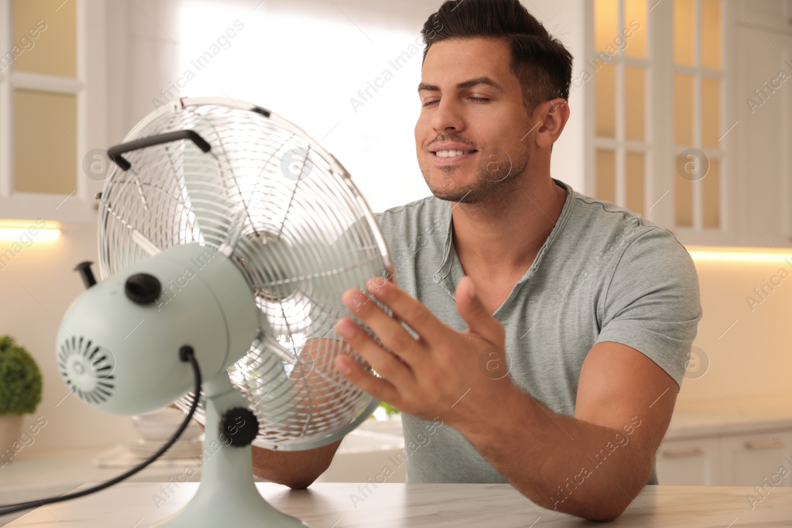 Photo of Man enjoying air flow from fan at table in kitchen. Summer heat