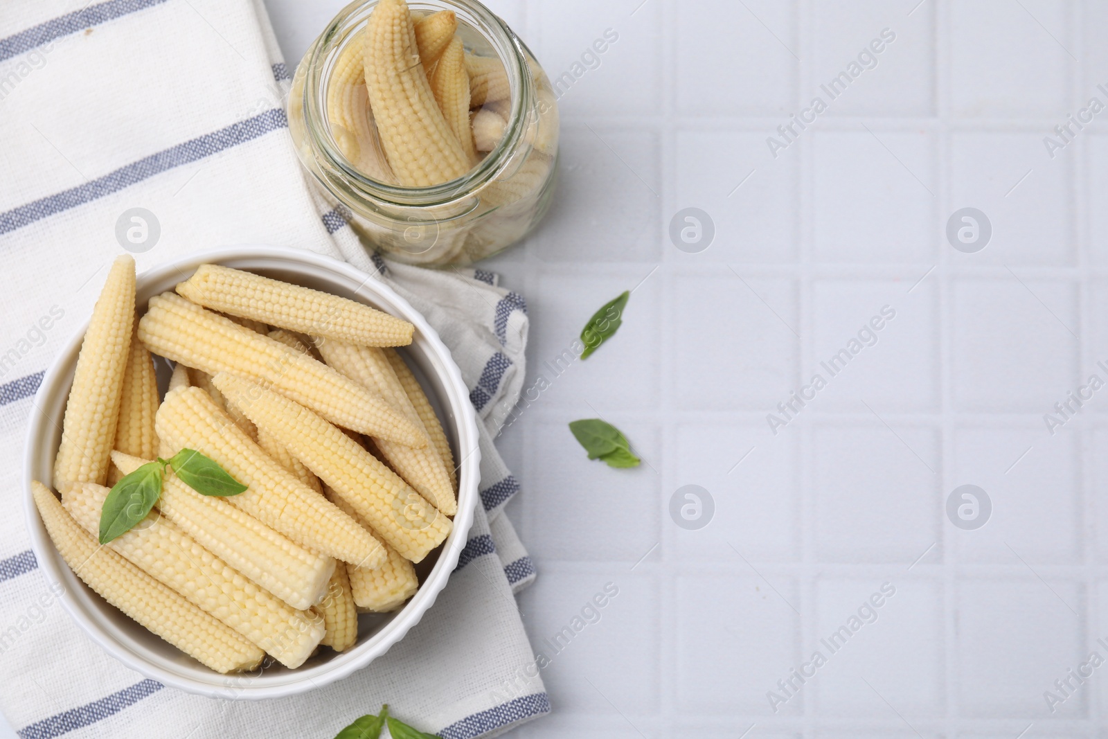 Photo of Canned baby corns with basil on white tiled table, flat lay. Space for text