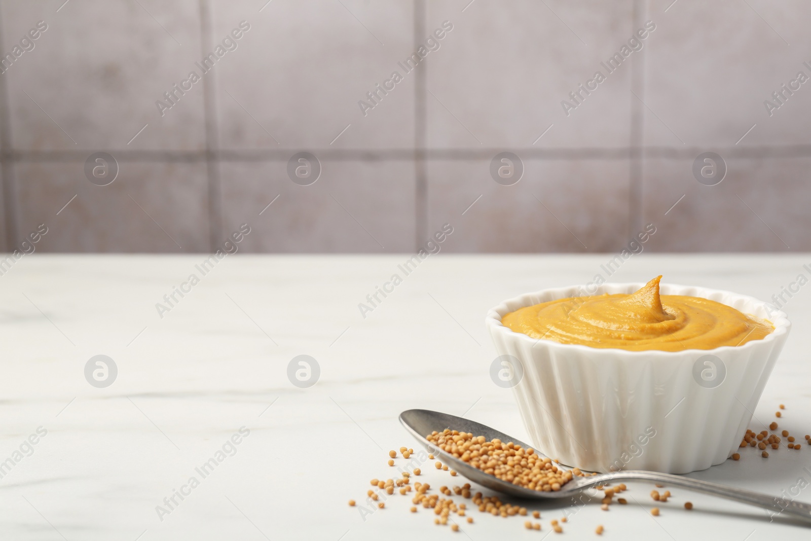 Photo of Tasty mustard sauce in bowl and spoon with dry seeds on white marble table, space for text