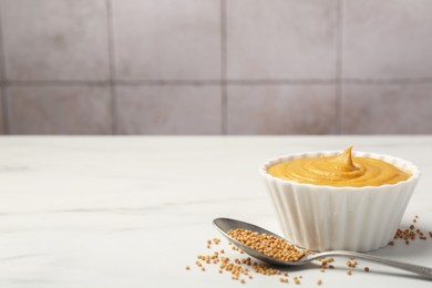 Photo of Tasty mustard sauce in bowl and spoon with dry seeds on white marble table, space for text